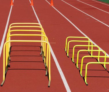 A red track set up for speed and agility work with different sized plastic yellow hurdles in lanes and orance construction cones at the end of the zone.