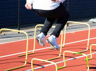 Side rear iw of a young high school athlete is boundind over small yellow banana hurdles on a red track for strength and agility practice.