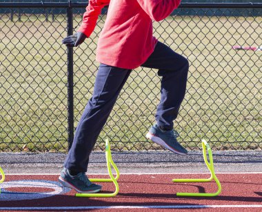 Side view of a high school boy standing over a yellow mini hurdle in the track A-Position on a cold but sunny day at track and field practice.