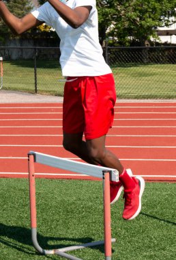 Güneşli bir bahar günü pistte saha antrenmanı sırasında yeşil bir çayırda engelin üzerinden atlayan Afrikalı Amerikalı bir sporcunun dikey görüntüsü..