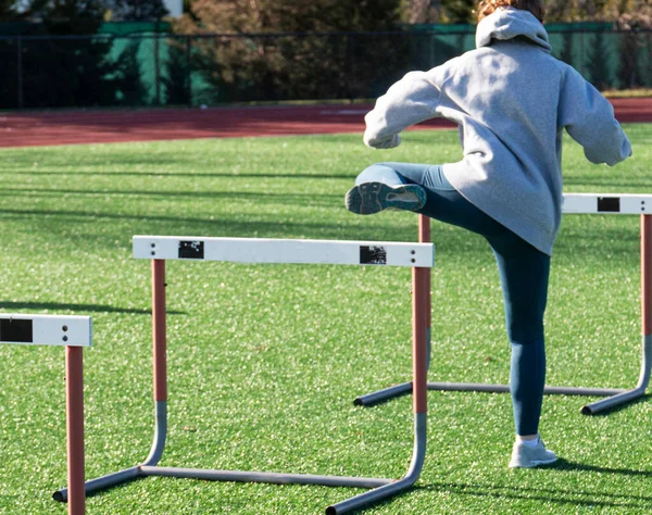 Rear view of a high school girl running over hurdles with her trail leg doing training drills on a green turf field.