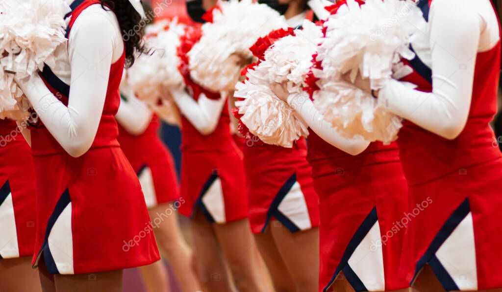 High school cheerleaders cheering at a basketball game indoors holding white and red pompoms.