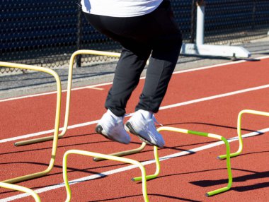 Liseli bir çocuk, çeviklik antrenmanı sırasında kırmızı bir pistte küçük sarı engellerin üzerinden atlıyor..