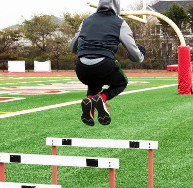 Kış aylarında güç ve çeviklik eğitimi sırasında çim sahasında yüksek engellerden atlayan liseli bir çocuğun arka görüntüsü..