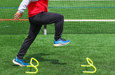 Pistte bir liseli koşucunun yan görüntüsü. A pozisyonu, spor antrenmanı yaparken küçük sarı bir engelin üzerinde..