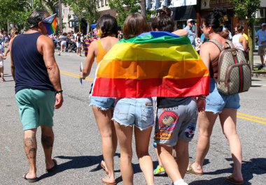 Babil, New York, ABD - 5 Haziran 2022: Babylon Village Pride Car Parade 2022 'nin başlamasını bekleyen eşcinsel bir gurur bayrağına sarılı olarak sokakta yürüyen insanların görüntüsü