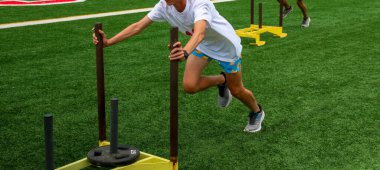 Runners pushing sleds with weights on a turf field during strength and agility drills at a high school summer camp.