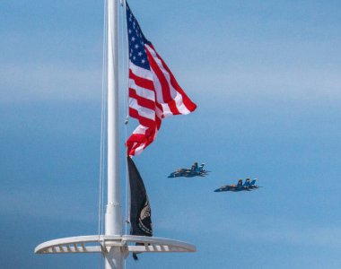 U.S. Navy Blue Angels flying in formation behind an American Flag blowing in the wind during an airshow.