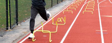 A high school runner trips over yellow mini hurdles set up on a track to run over during the wicket drill at track practice.