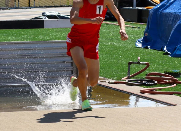 A high school runner wearing the number one on his red uniform exiting the steeplechase water pit with a little but of splashing during a race.