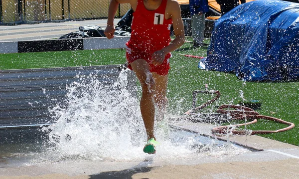 A high school boy splashing as he exits the steeplechase water jump during a race that he is winning.