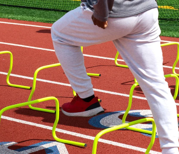 A high school track and field runner performing speed and agility drills over yellow mini hurdles on a track.