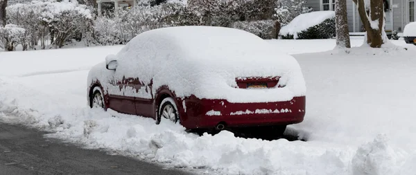A red car covered in snow parked in the street during a storm so the snowplows had to go around and block the car in.