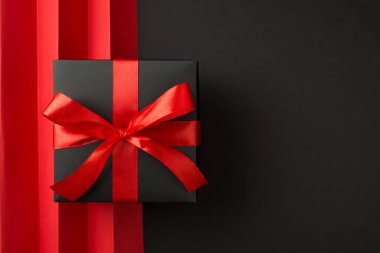 Kırmızı kurdeleli siyah hediye kutusunun üst görünümü ve izole edilmiş siyah zemin üzerinde dikey kıvrımlı ve boş arkaplan ile kırmızı sayfa