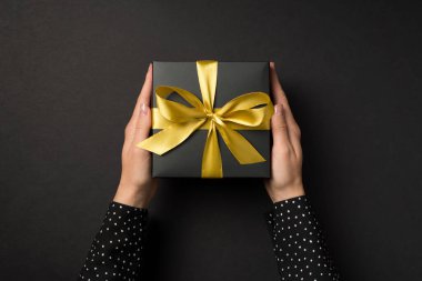 Üzerinde altın saten kurdele olan siyah bir hediye kutusu taşıyan polka noktalı tişörtlü ellerin izole edilmiş siyah arka planda olduğu ilk kişi fotoğrafı.