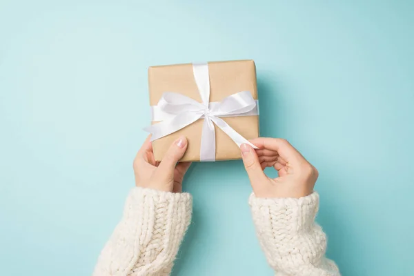 First person top view photo of woman's hands in white cozy sweater unpacking craft paper giftbox with white satin ribbon bow on isolated pastel blue background
