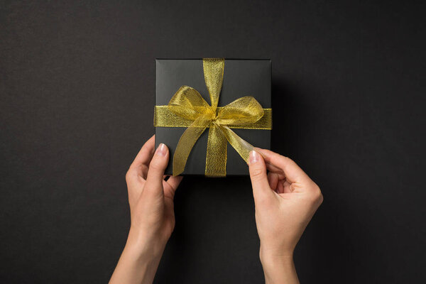 First person top view photo of hands unpacking black giftbox with golden mesh ribbon bow on isolated black background