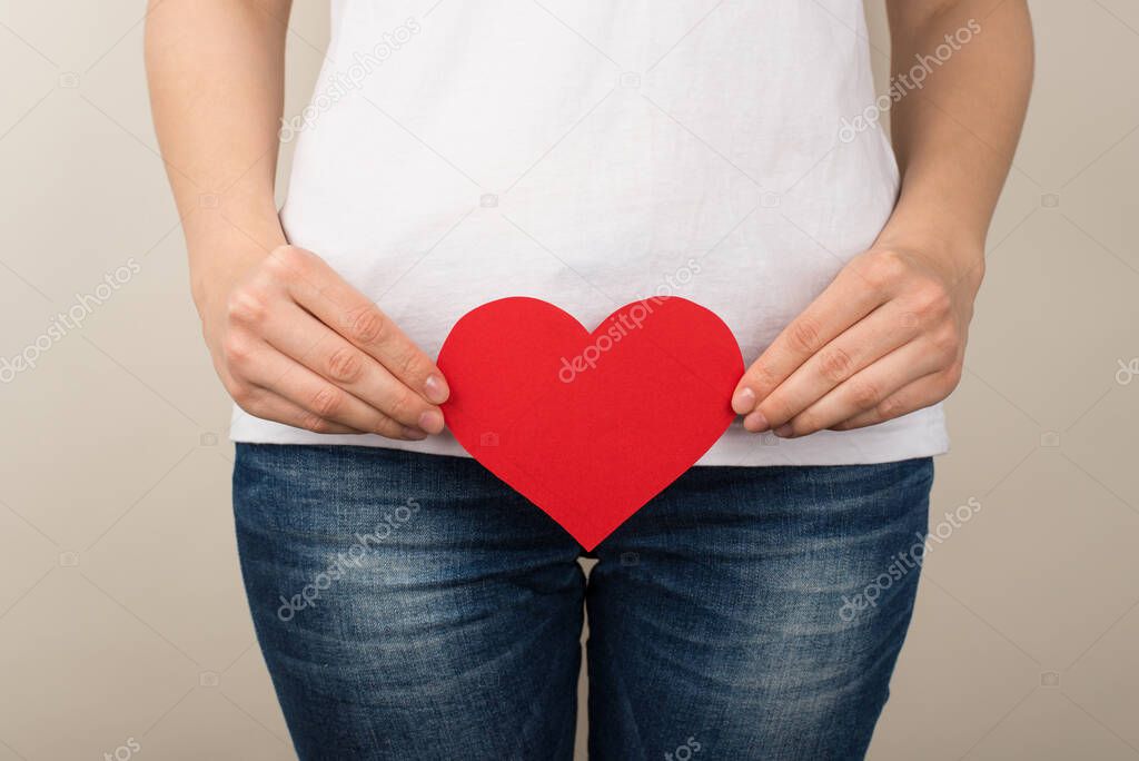 Cropped closeup photo of woman in white t-shirt and jeans holding red paper heart near crotch on isolated grey background