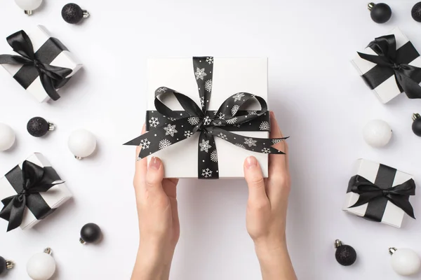 白と黒のクリスマスの装飾の上に黒いサテンリボン弓と白いギフトボックスを与える女性の手の最初の人のトップビューの写真孤立した白の背景にプレゼントとボール — ストック写真