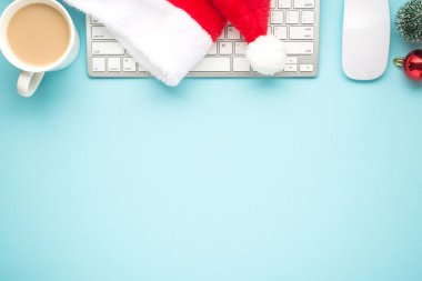 Noel Baba şapkasının üst görüntüsü klavyede bilgisayar faresi bardağı sıcak çam ağacı oyuncağı ve izole edilmiş pastel mavi arka planda kırmızı noel ağacı topu.