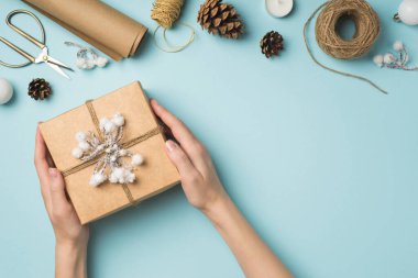 Üstten bakıldığında, kar dalı ve sicim Noel ağacı ile süslenmiş Kraft kağıt hediye kutusu tutan ellerin fotoğrafı izole edilmiş pastel mavi arka planda el işi aletler, boş alan.