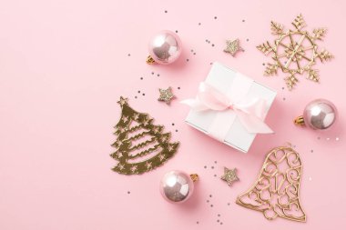 Pembe noel süslemelerinin en üst görüntüsü. Parlayan yıldızlar, altın çan çanı, kar tanesi şekilli süsler ve izole edilmiş pastel pembe arka planda pembe fiyonklu beyaz hediye kutusu.