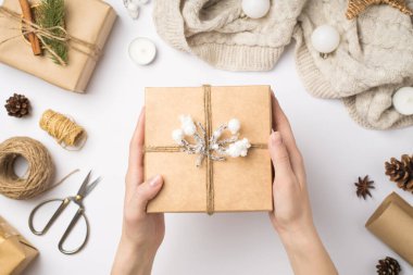 First person top view photo of hands holding craft paper giftbox decorated with twine and snow branch over sweater christmas tree balls and handicraft tools on isolated white background clipart