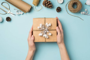 Üstten bakan ilk fotoğrafta kadın eli, kar dalı ile süslenmiş hediye kutusunu topların üzerinde tutuyordu. Makas makaralar, iplik rulo kağıt mum makaraları ve izole edilmiş pastel mavi arka plan üzerindeki koniler.