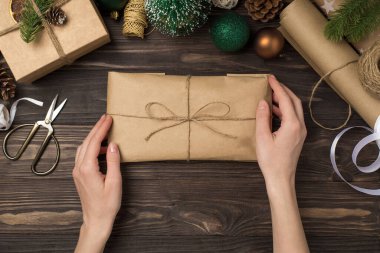 Üstten bakıldığında, kadınların el sanatları el işi kağıt paketini tutarken çekilmiş ilk fotoğrafta sicim yay yay, yeşil altın toplar, çam dalları ve el işi aletler izole edilmiş, ahşap masa arkasında.