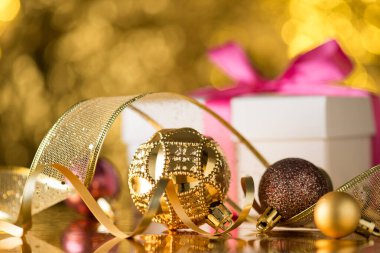 Altın ve kahverengi Noel ağacı süslemelerinin yakın plan fotoğrafı topları yılanbalığı kurdelesi ve parlak altın renkli arka planda pembe fiyonklu beyaz hediye kutusu.