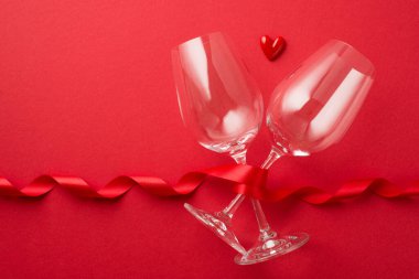 Sevgililer Günü dekorasyonunun en üst görüntüsü kırmızı kıvrımlı kurdele ve iki şarap kadehi arasında kopyalanmış kırmızı arka planda küçük kalp