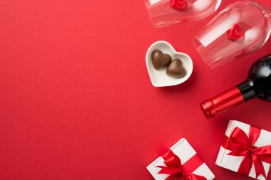 Sevgililer Günü süslemelerinin en üst görüntüsü kırmızı kurdeleli beyaz hediye kutuları iki şarap kadehinde küçük kalpler şekerlemeli fincan tabağı ve izole edilmiş kırmızı arka planda şarap şişesi.