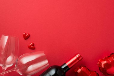 Sevgililer Günü süslemelerinin en üst görüntüsü. İki şarap kadehi, kalp şeklinde balonlar ve izole edilmiş kırmızı arka planda boş uzay boşluğu olan şarap şişesi arasında küçük kalpler.