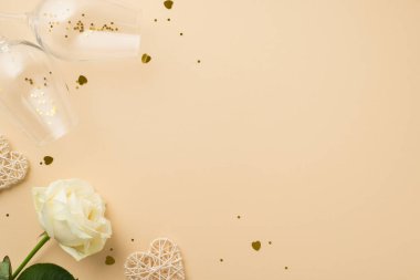 Beyaz gülün üst görüntüsü altın pullu iki şarap kadehi kalp şeklinde konfeti ve izole edilmiş bej arkaplanlı rattan kalpler.