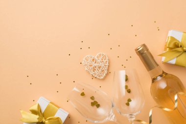 Sevgililer Günü dekorunun en üst görüntüsü altın fiyonklu beyaz hediye kutuları, şarap şişesi, rattan kalp şarap kadehleri, kalp şeklinde konfeti ve izole edilmiş pastel bej arka planda pullar.