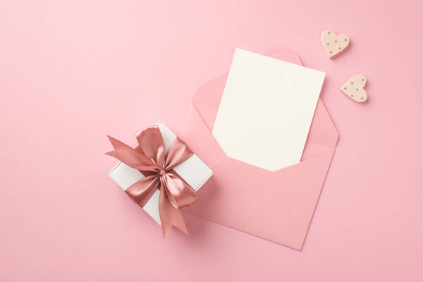 Top view photo of white giftbox with pink satin ribbon bow decorative hearts and open pink envelope with paper sheet on isolated pastel pink background with copyspace