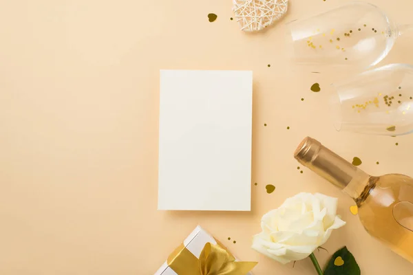 Top view photo of paper sheet white giftbox with gold bow two wineglasses wine bottle sequins white rose and heart shaped golden confetti on isolated beige background with blank space