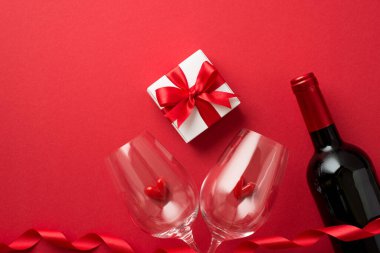 Sevgililer günü süslemelerinin en üst görüntüsü kırmızı kurdeleli hediye kutusu iki şarap kadehinde küçük kalpler ve izole edilmiş kırmızı arka planda boş uzay boşluğu olan şarap şişesi