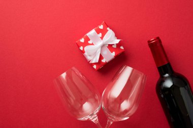 Sevgililer Günü süslemelerinin en üst görüntüsü kırmızı ambalaj kağıdıyla kaplanmış hediye kutusu. Kupa desenleri ve beyaz kurdele ile izole edilmiş kırmızı arka planda iki şarap kadehi ve şarap şişesi.