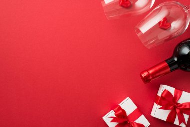 Sevgililer Günü süslemelerinin en üst görüntüsü kırmızı kurdeleli beyaz hediye kutuları izole edilmiş kırmızı arka planda iki şarap kadehi ve şarap şişesinde küçük kalpler var.