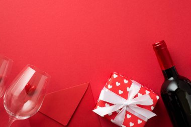 Kırmızı ambalaj kağıdıyla hediye kutusunun en üst görüntüsü kalpler deseni ve beyaz kurdele küçük kalpler iki şarap kadehinde kırmızı zarf ve izole edilmiş kırmızı arka planda şarap şişesi