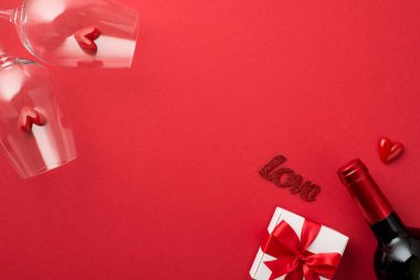 Sevgililer Günü süslemelerinin en üst görüntüsü kırmızı kurdeleli beyaz hediye kutusu iki şarap kadehinde küçük kalpler izole edilmiş kırmızı arka planda aşk ve şarap şişesi ve boş alan.