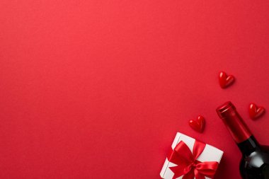Sevgililer Günü süslemelerinin üst görüntüsü kırmızı kurdeleli beyaz hediye kutusu izole edilmiş kırmızı arka planda küçük kalpler ve şarap şişesi boş alan