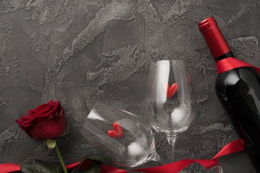 Sevgililer Günü süslemelerinin en üst görüntüsü küçük kalpli iki şarap kadehi izole edilmiş koyu gri beton arka planda kırmızı gül kırmızı kurdele ve şarap şişesi.