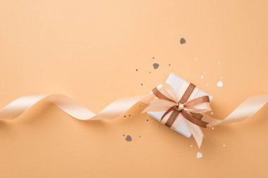 Sevgililer Günü dekorunun en üst görüntüsü gümüş kalp şeklinde konfeti şekilli konfeti parıltılı beyaz hediye kutusu, izole edilmiş bej arka planda parlak kahverengi ve açık bej kıvrımlı kurdele.