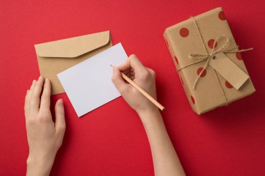 Sevgililer Günü dekorasyonunun birinci şahıs fotoğrafı. Kadın elleri, polka desenli zarf hediye kutusu ve kopyalanmış kırmızı arka planda sicim yayı yazıyor.