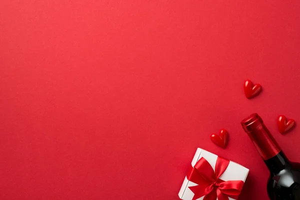 Top view photo of valentine's day decorations white giftbox with red bow small hearts and wine bottle on isolated red background with blank space