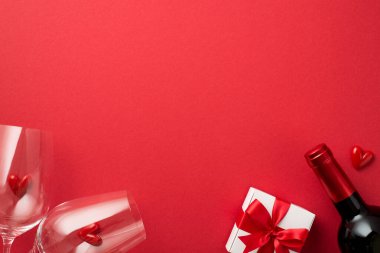 Top view photo of valentine's day decorations white giftbox with red bow small hearts in two wineglasses and wine bottle on isolated red background with blank space