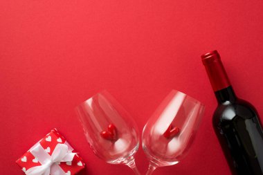 Top view photo of valentine's day decorations giftbox in red wrapping paper with pattern of hearts and bow small hearts in two wineglasses and wine bottle on isolated red background with copyspace