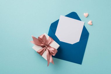 Top view photo of open blue envelope with letter small pink hearts and white giftbox with light pink ribbon bow on isolated pastel blue background with empty space